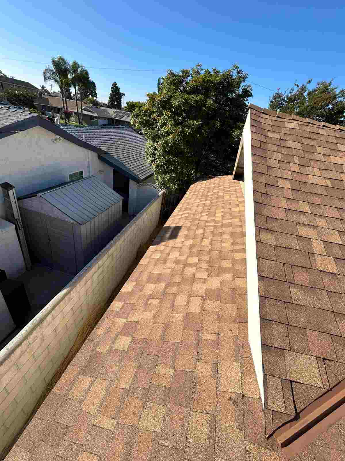 Brown shingle slopes beside a taller roof section and a white vertical junction, with a block wall and adjacent buildings