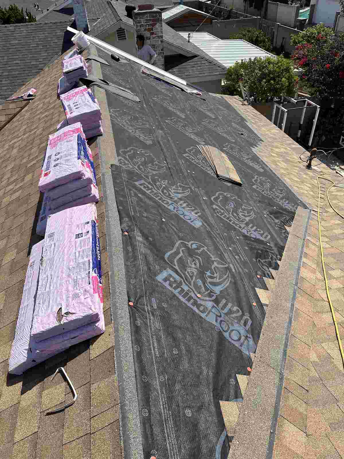 Dark printed underlayment rolled across a roof with pink wrapped roofing bundles and exposed deck and shingles nearby